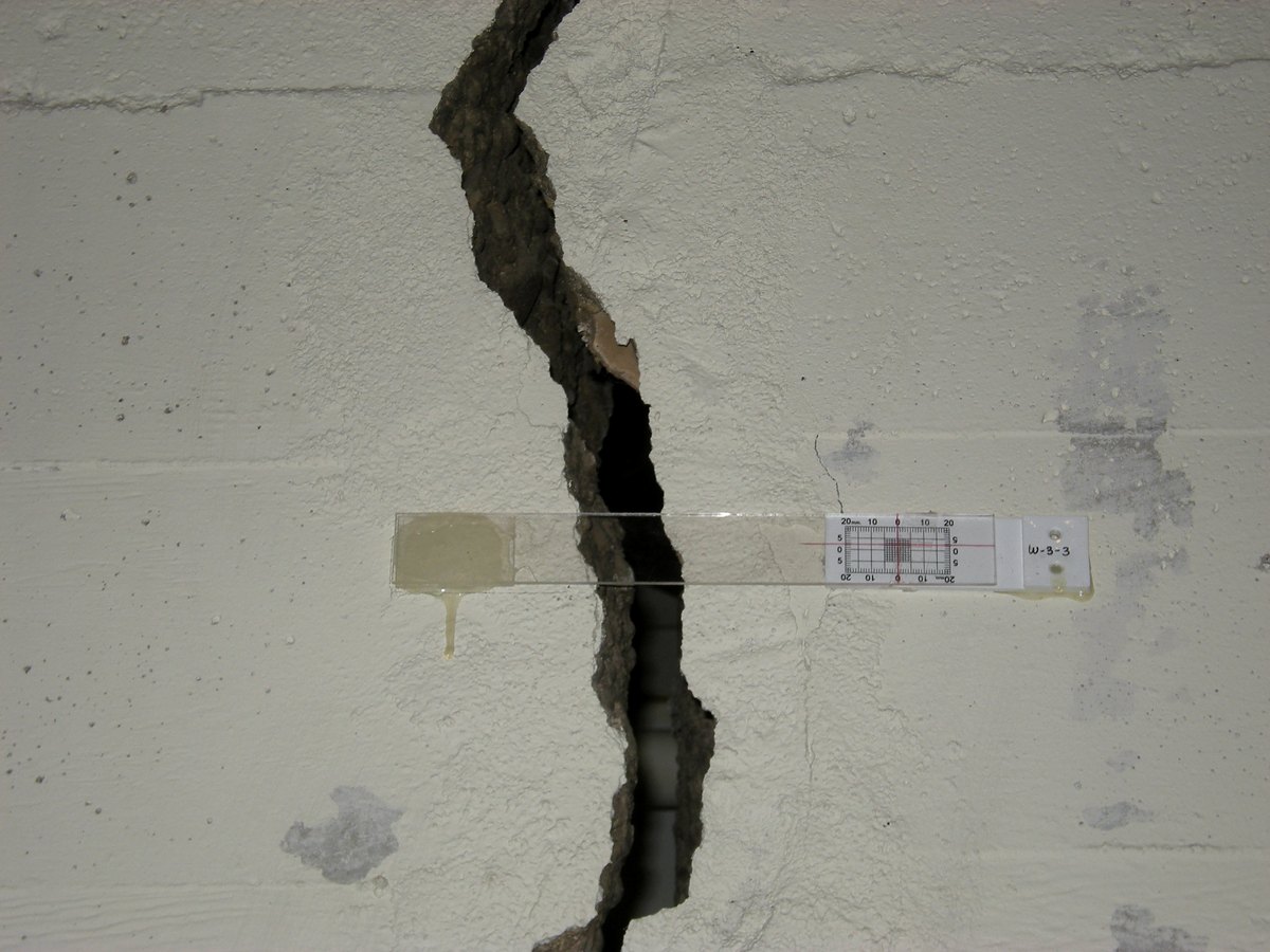 Grieta ancha y de trayectoria irregular en un muro interior, con un testigo de escala y una banda de yeso atravesándola